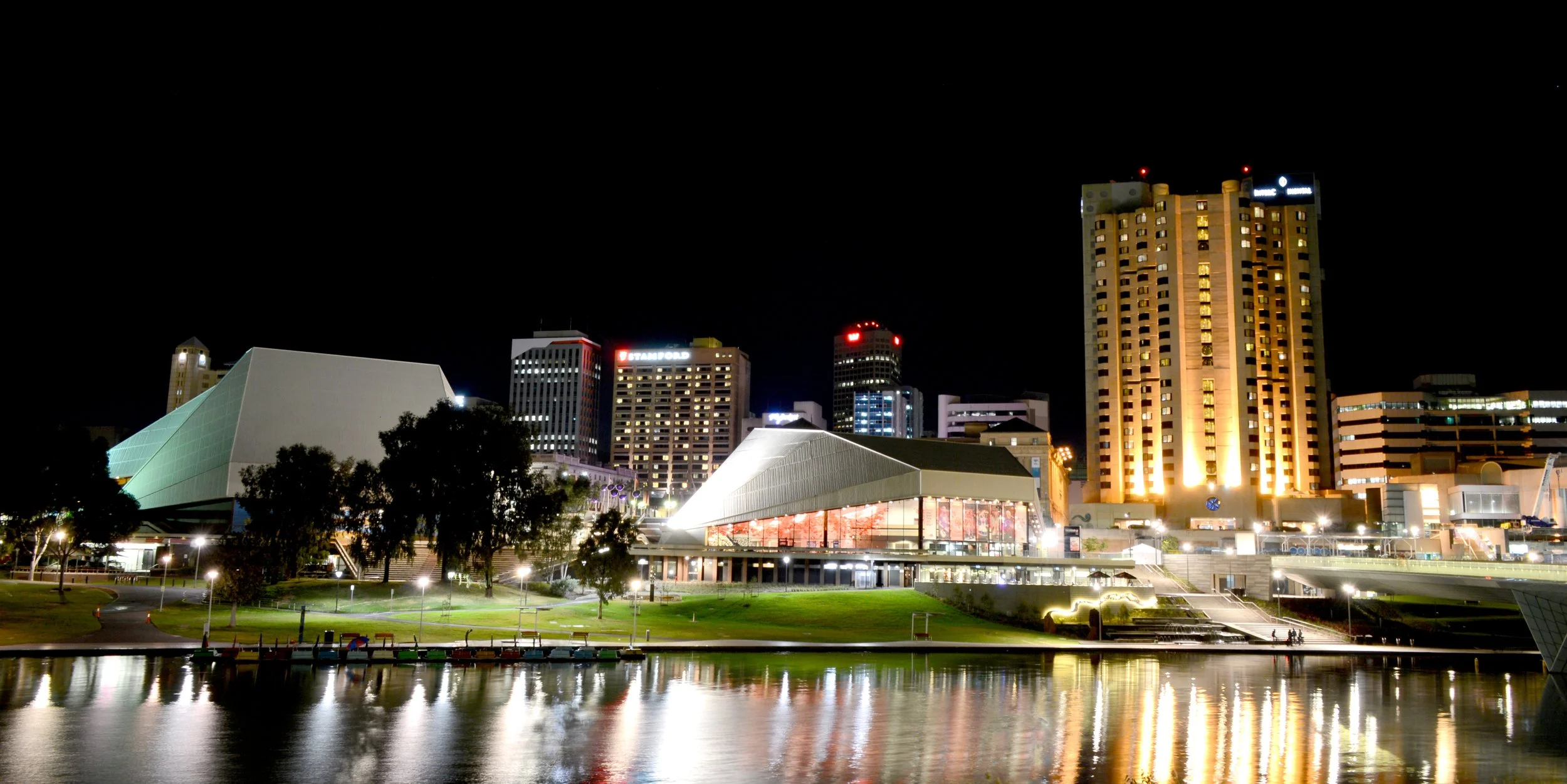 Night view of a city skyline with lit buildings, modern architecture, trees, and a waterfront reflecting city lights.
