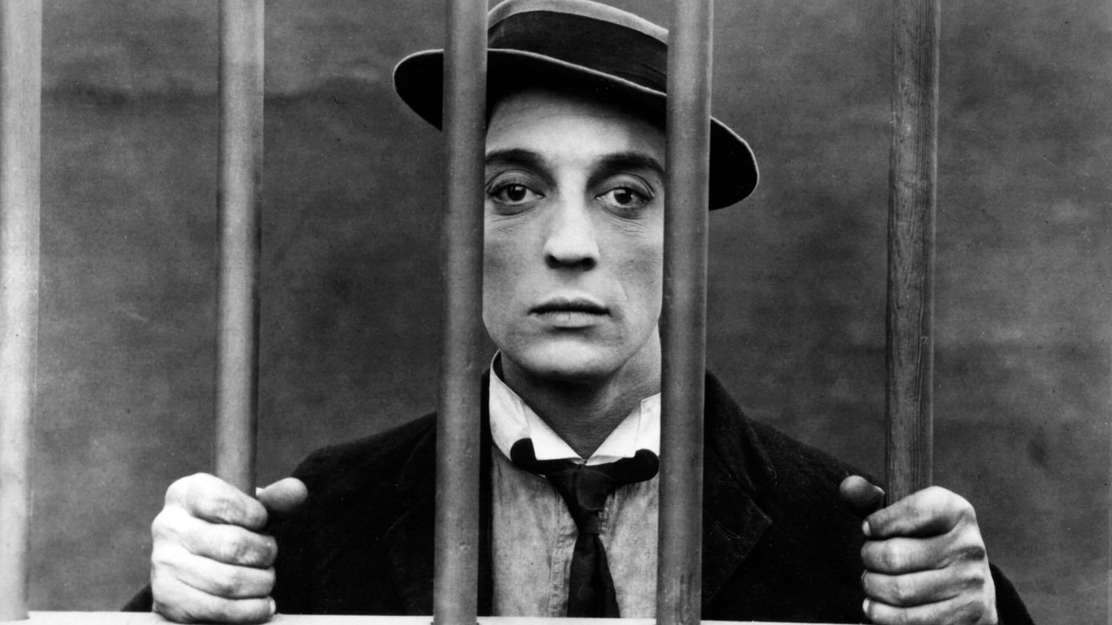 A black and white photograph of a person behind bars, holding the bars with both hands. The person is wearing a hat, a white shirt, and a dark tie, and has a serious expression on their face.