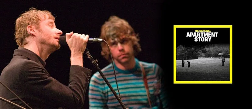 Two young men on stage, one singing into a microphone with eyes closed, the other standing in the background wearing a striped shirt, with a black-and-white cover of 'The National Apartment Story' album in the background.