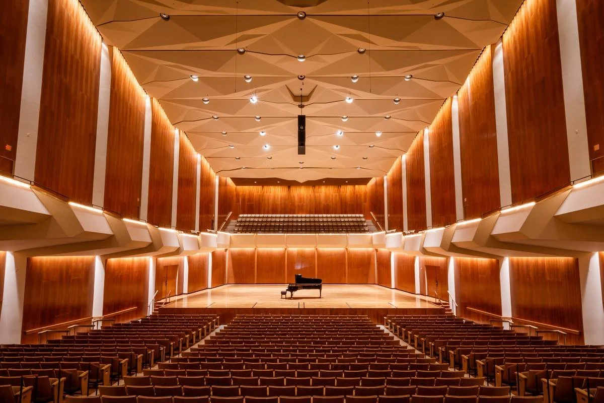 An empty concert hall with a grand piano on stage and carpeted seating area in front.