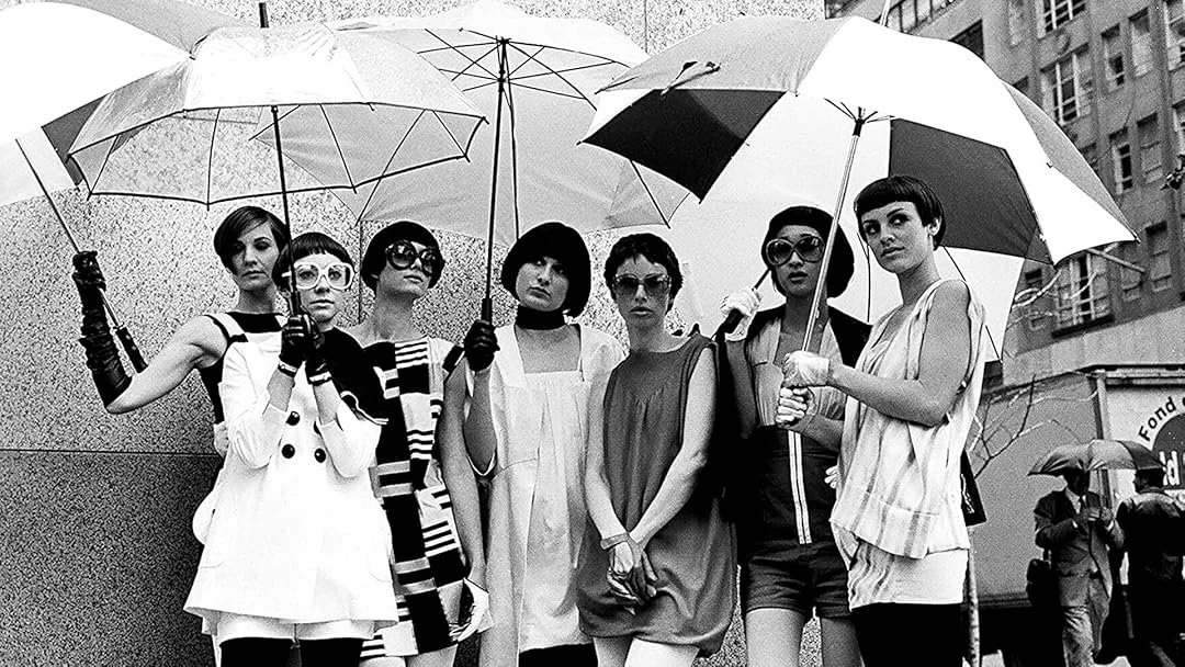 Group of seven women standing outdoors on a city street, holding umbrellas in the rain. They are dressed in stylish, vintage-inspired outfits and appear to be posing for a photograph.