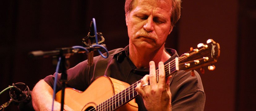 A man with reddish hair and mustache playing an acoustic guitar in a recording studio.