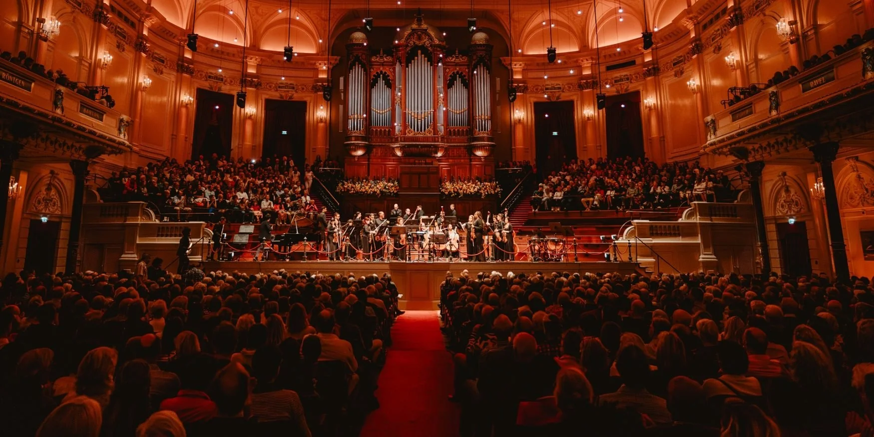 Orchestra performing on stage in a grand, ornately decorated concert hall filled with an audience.