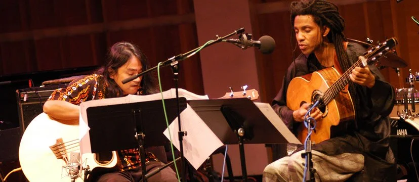 Two musicians, one playing an acoustic guitar and the other playing a classical guitar, are performing in a recording studio.
