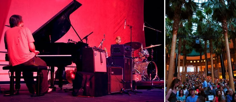 Musicians performing on stage with a red backdrop, including a pianist, drummer, and other instruments, with an audience seated outdoors under palm trees in a lit venue.