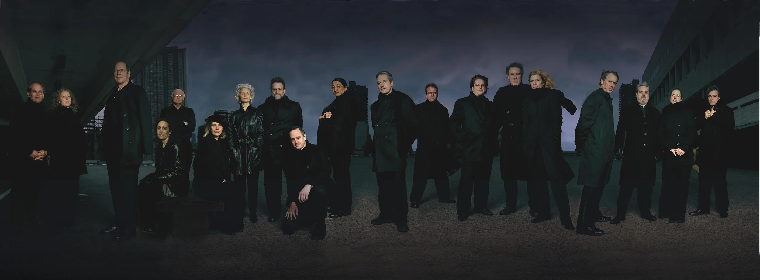A group of people dressed in black standing and sitting under a dark, cloudy sky in an urban setting.