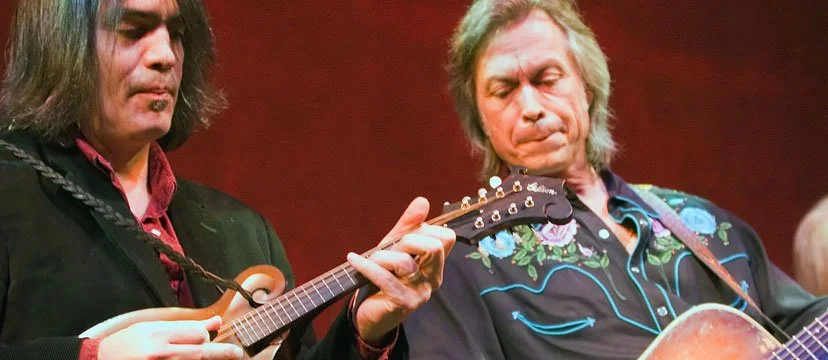 Two men playing guitars, one appears to be strumming and the other looking down, on stage with a red background.