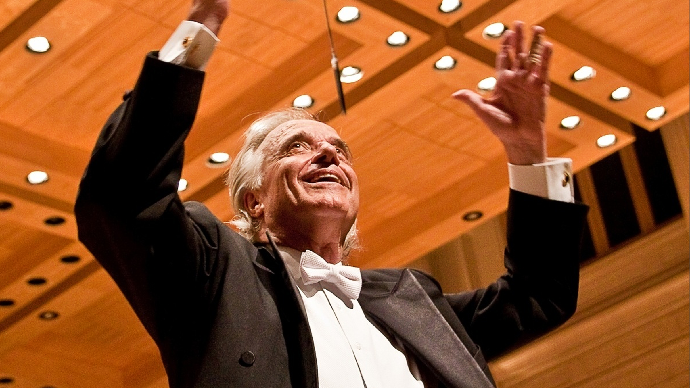 An elderly man in a tuxedo with a white bow tie conducting an orchestra, with his arms raised and a joyful expression on his face, set against a wood-paneled ceiling with circular lights.