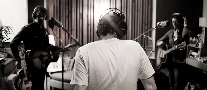 A person wearing headphones recording in a studio with two musicians playing guitars on either side.