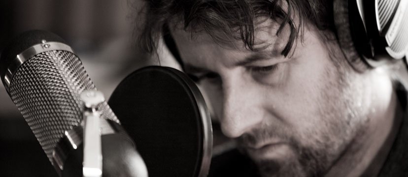 A man with headphones singing or speaking into a studio microphone with a pop filter.