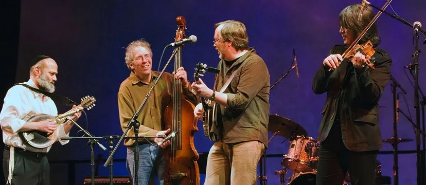 Four musicians performing on stage, playing guitar, upright bass, and violin, with microphones and drum set in the background.