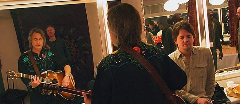 A person in the foreground is playing guitar with two men listening; one man in the background is also playing guitar, and the other man is sitting with his eyes closed, possibly singing or listening to music.