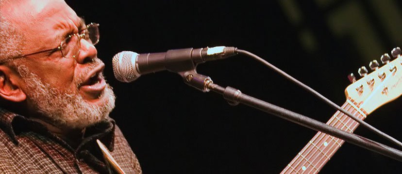 An elderly man singing into a microphone while playing an electric guitar.