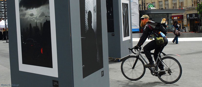 A man in black cycling gear, with a backpack and cap, riding a black bicycle past an outdoor art display or advertisement panels in an urban city environment. Pedestrians walk in the background near storefronts.