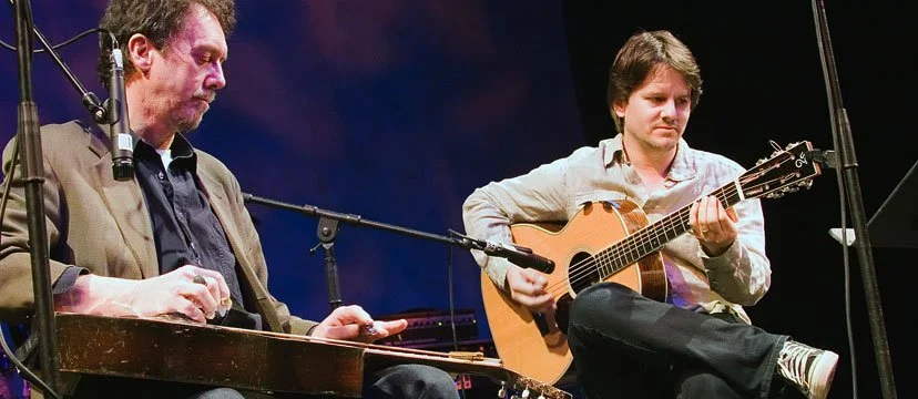 Two male musicians performing on stage, one playing a lap steel guitar and the other playing an acoustic guitar.
