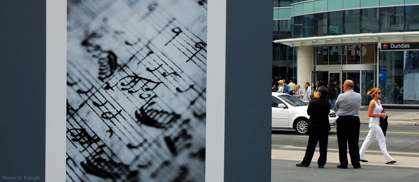 A large vertical outdoor billboard displays a close-up of sheet music, with people standing on the sidewalk near a modern building with a 'Dundas' sign across the street.