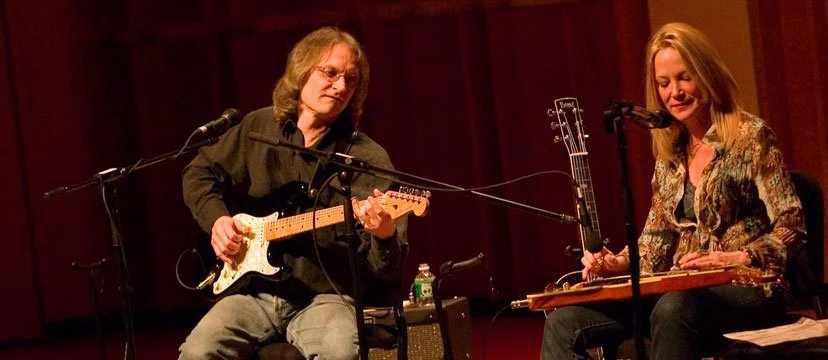 Two women playing guitars on a stage with microphones, one with an electric guitar and the other with a lap steel guitar, in a dimly lit performance setting.