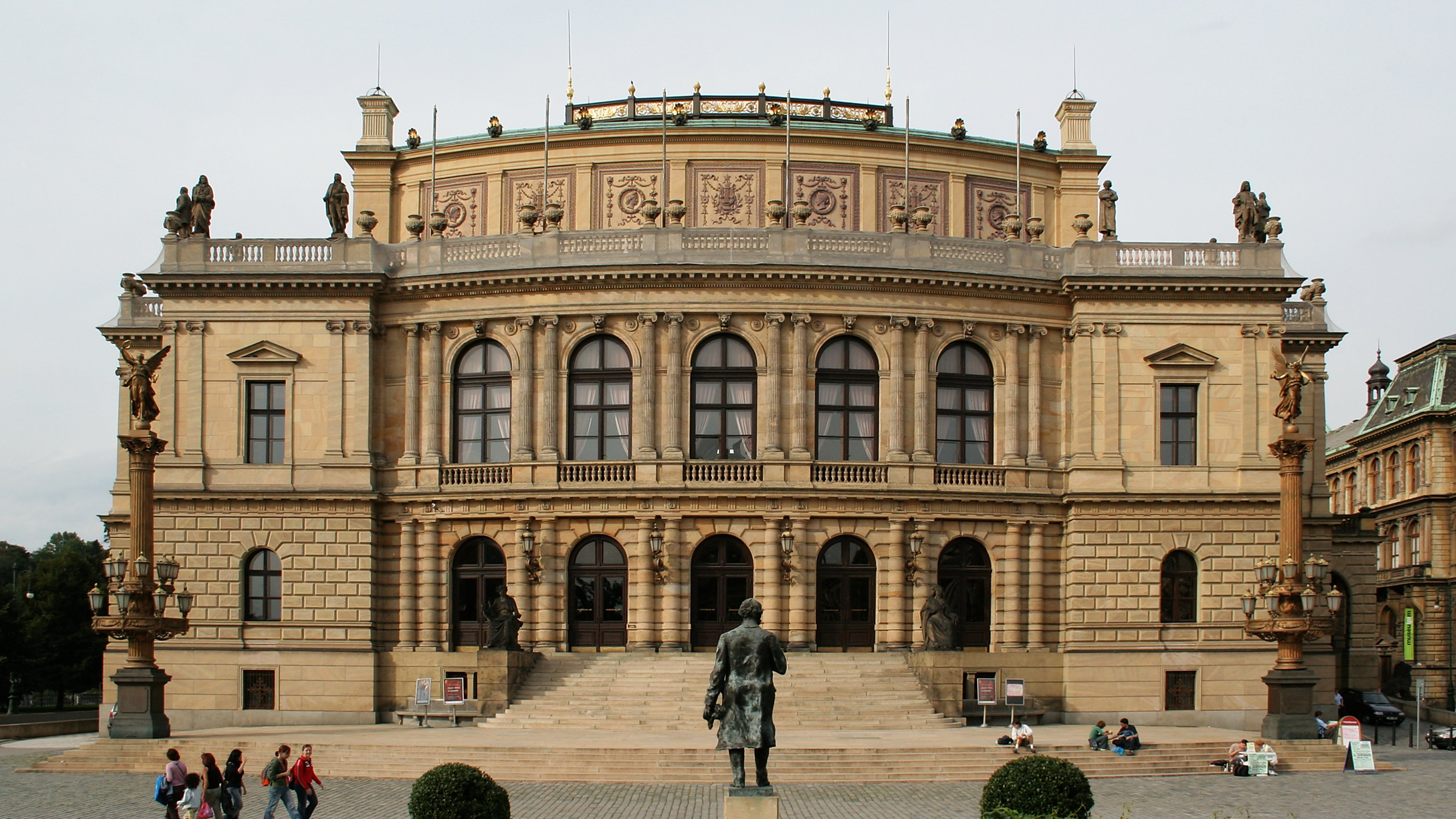 A grand, historic building with ornate architecture, statues on the roof, and a staircase leading to the entrance, with a bronze statue of a man in front and groups of people sitting and walking around.