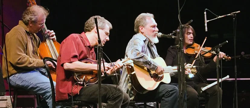 Four musicians performing on stage, playing guitars and violins, with microphones set up in front of them.