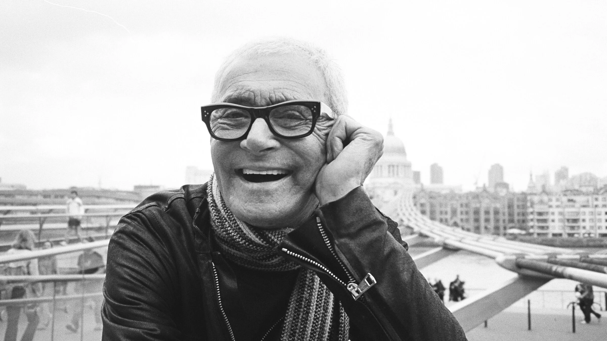 Smiling man wearing glasses and a leather jacket, sitting outdoors with a city skyline and a bridge in the background.