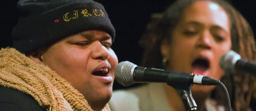 Two people singing into microphones, one wearing a beanie and scarf, with a blurred background.