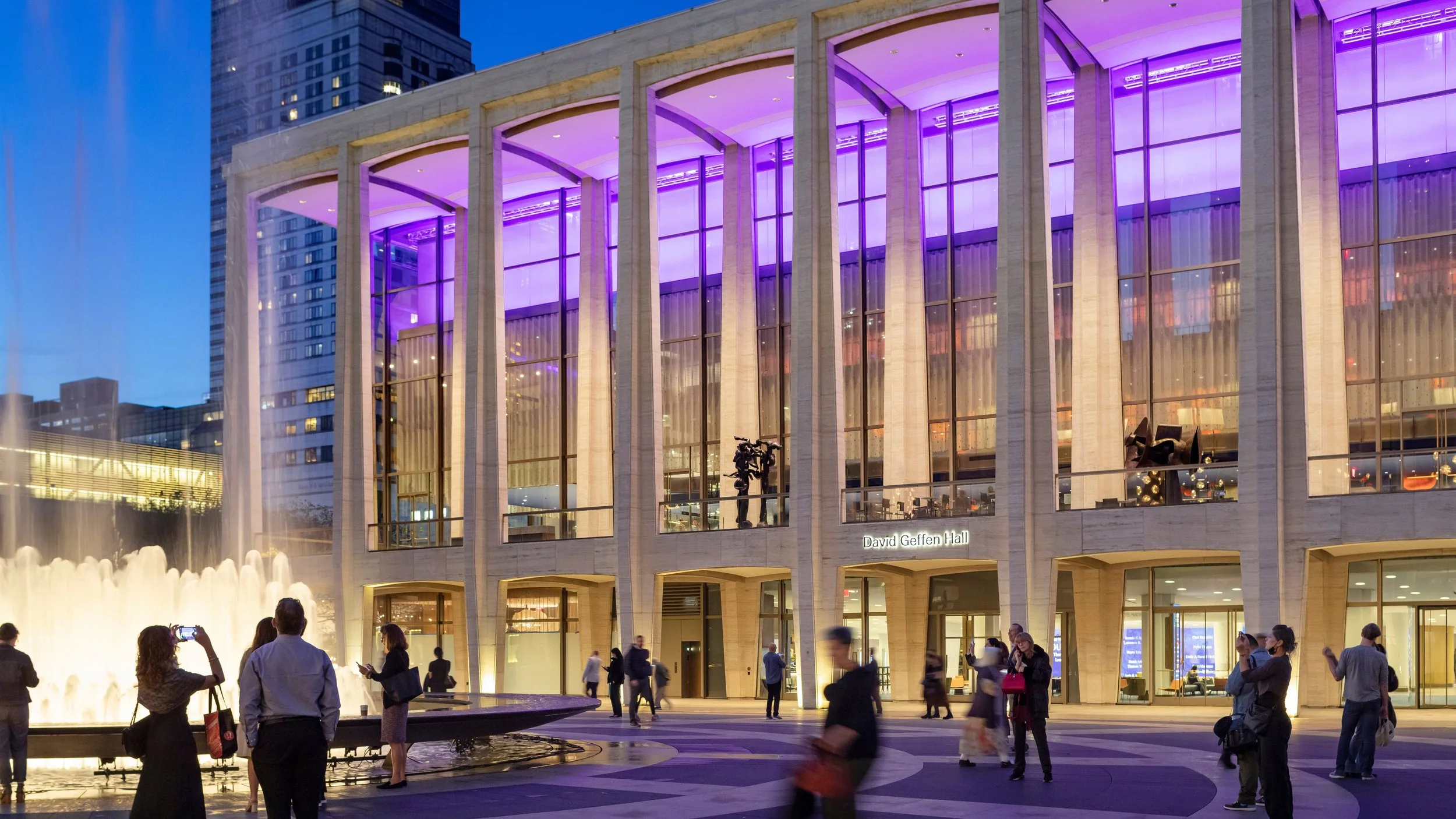 Nighttime view of David Geffen Hall with purple lighting on its modern glass and concrete exterior. People are gathered outside, some taking photos, near a fountain with water jets, in an urban setting with tall buildings in the background.