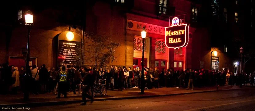 Long line of people waiting outside Massey Hall during evening, illuminated by streetlights and neon signs.