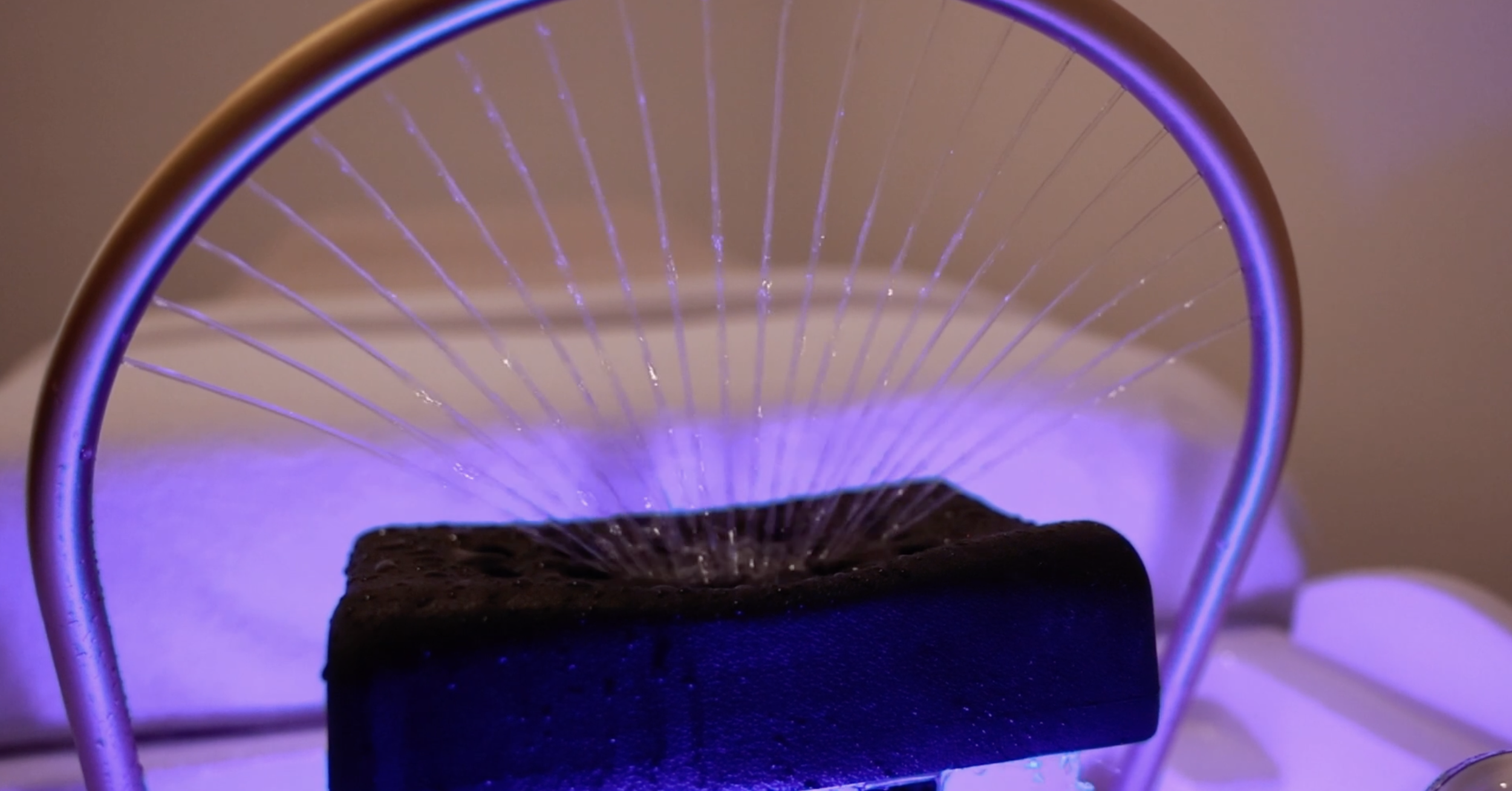 A close-up view of a black charcoal piece placed on a heat source, surrounded by a metallic structure with purple and blue lighting, likely part of a scientific or experimental setup.