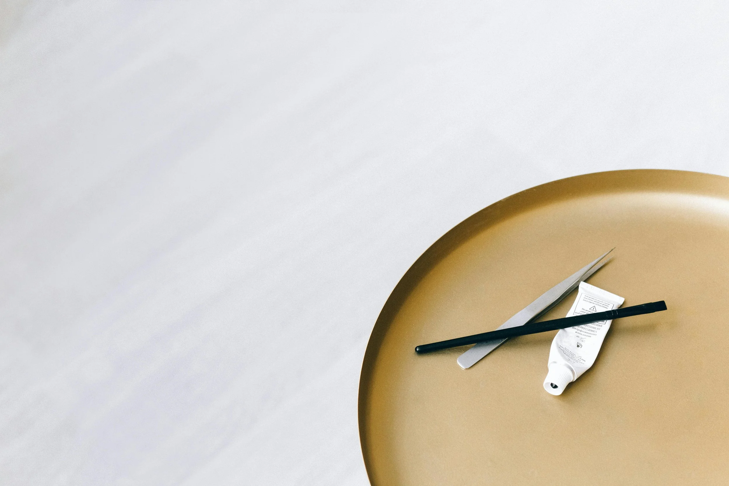 A round golden tray holding a pair of scissors, a black cosmetic applicator brush, and a white squeeze tube of cream or lotion on a plain white background.