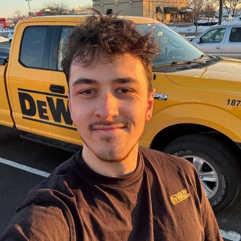 A young man with short, curly brown hair and a light beard taking a selfie in a parking lot during sunset. Behind him, there is a yellow pickup truck with 'DEWALT' branding on the side, with other vehicles and a building in the background.