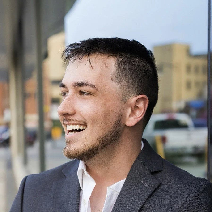A man in a suit smiling outdoors in an urban setting.