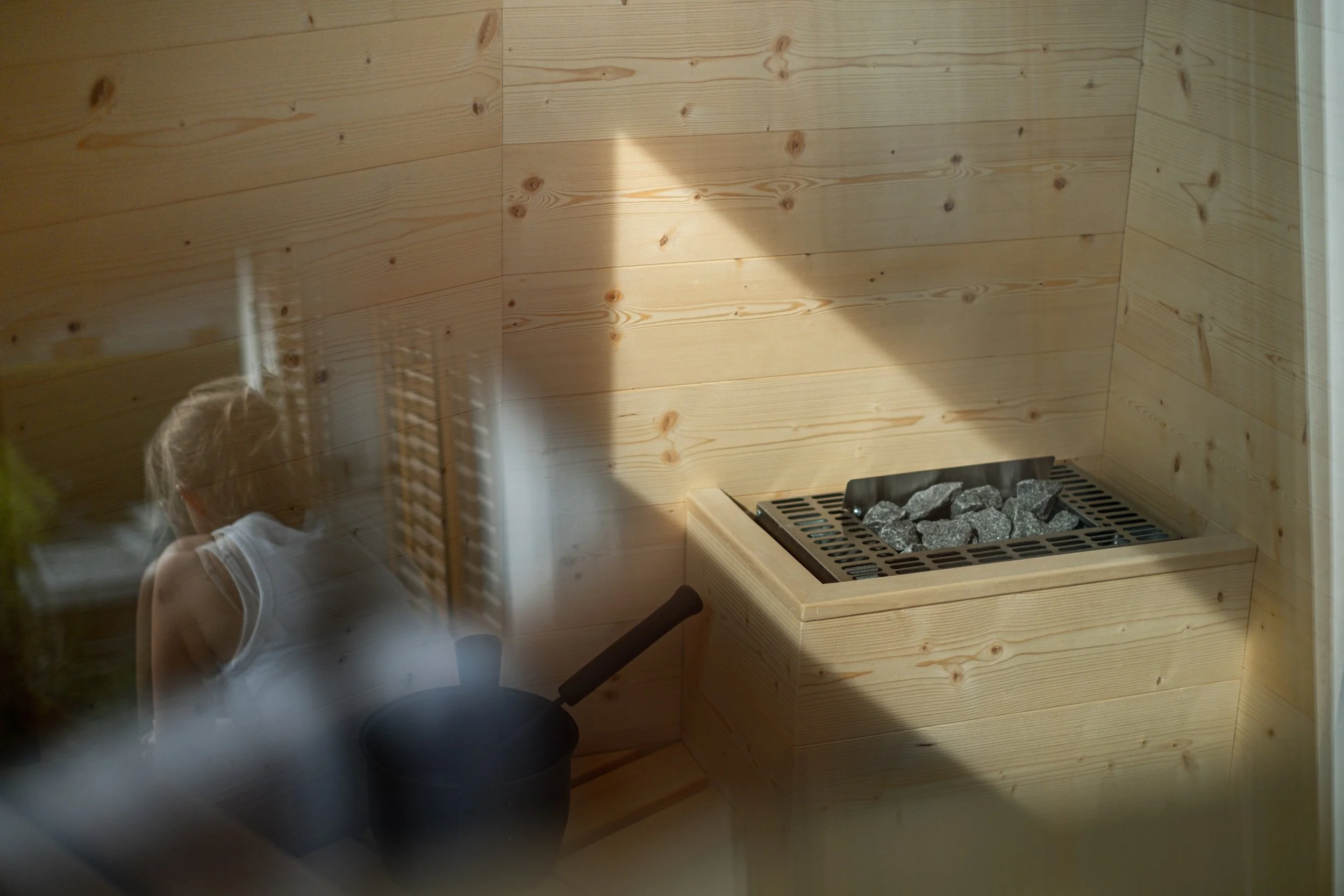 Innenansicht einer Sauna mit Holzverkleidung, einem Saunaofen mit Steinen und einer Stuhllehne, aufgenommen durch eine reflektierende Oberfläche.