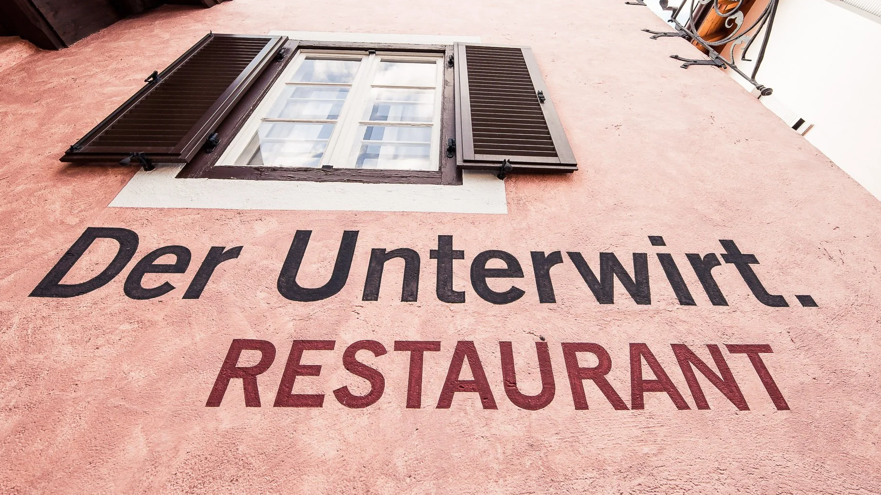 Ein Haus mit einer Wand, die pinkfarben ist. Es gibt eine Fenster mit braunen Fensterläden und die Worte "Der Unterwirt. RESTAURANT" an der Wand.