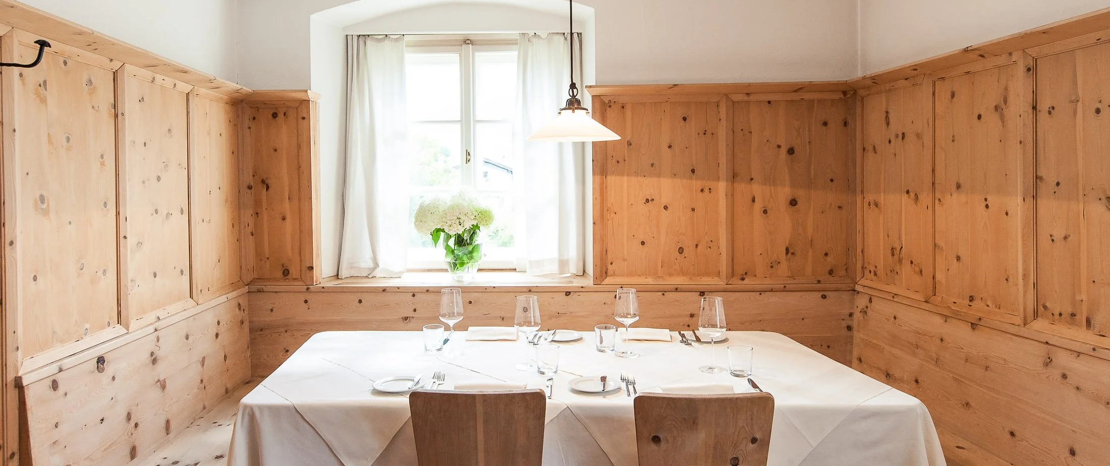 Ein Esszimmer mit holzgetäfelten Wänden, einem Tisch mit weißen Tischtuch, Geschirr und Gläsern, einer Fensterbank mit Vase und weißen Vorhängen.