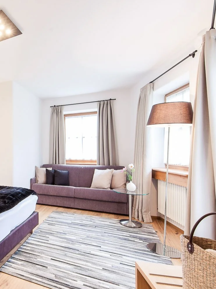 Wohnzimmer mit grauem Sofa, bunten Kissen, großem Fenster, Stehlampe, Vorhängen und Teppich