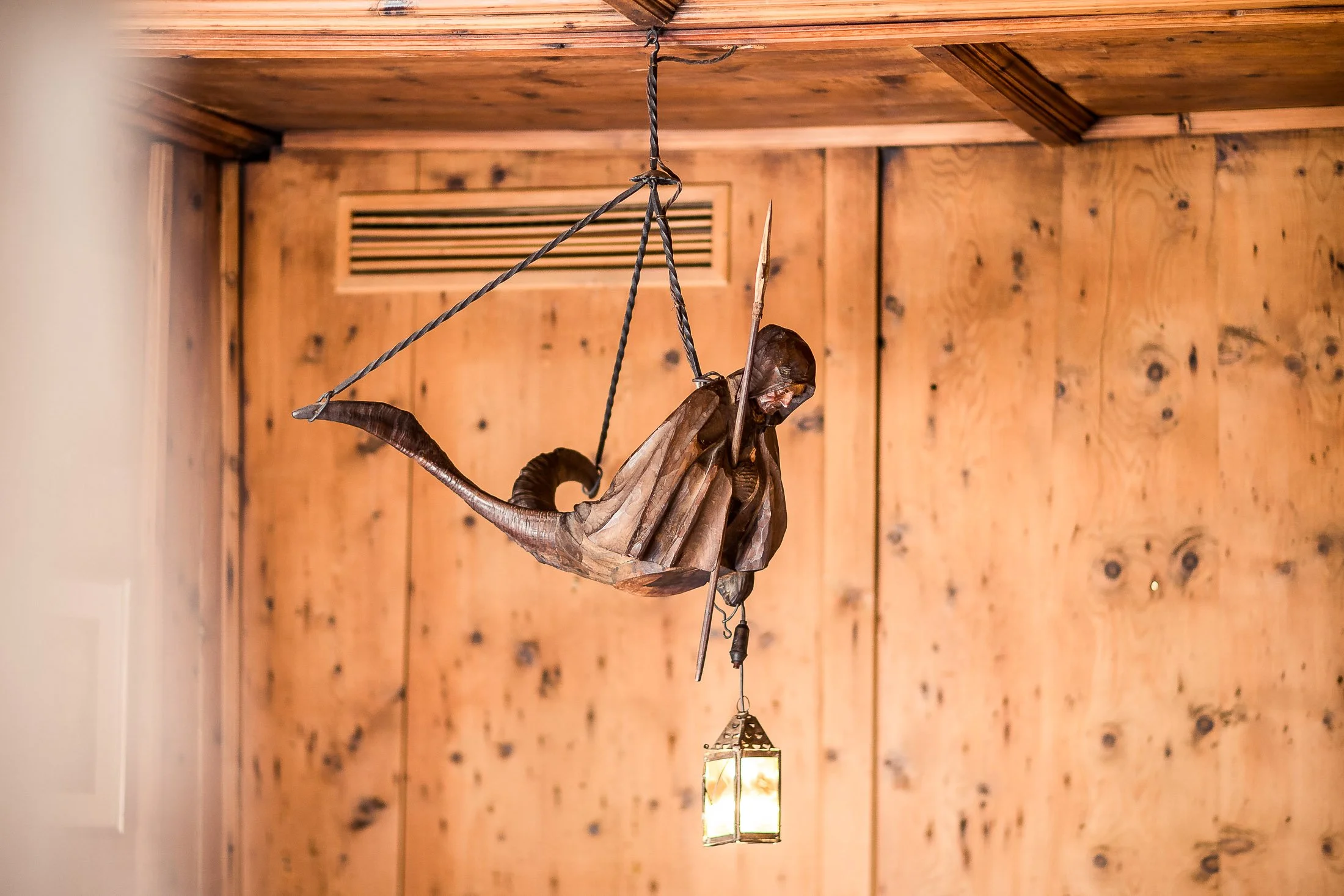Holzskulptur eines sitzenden Engels mit Flügeln und Heiligenschein, an einem Seil aufgehängt, mit laterngleichender Beleuchtung in einem Raum mit Holzvertäfelung.