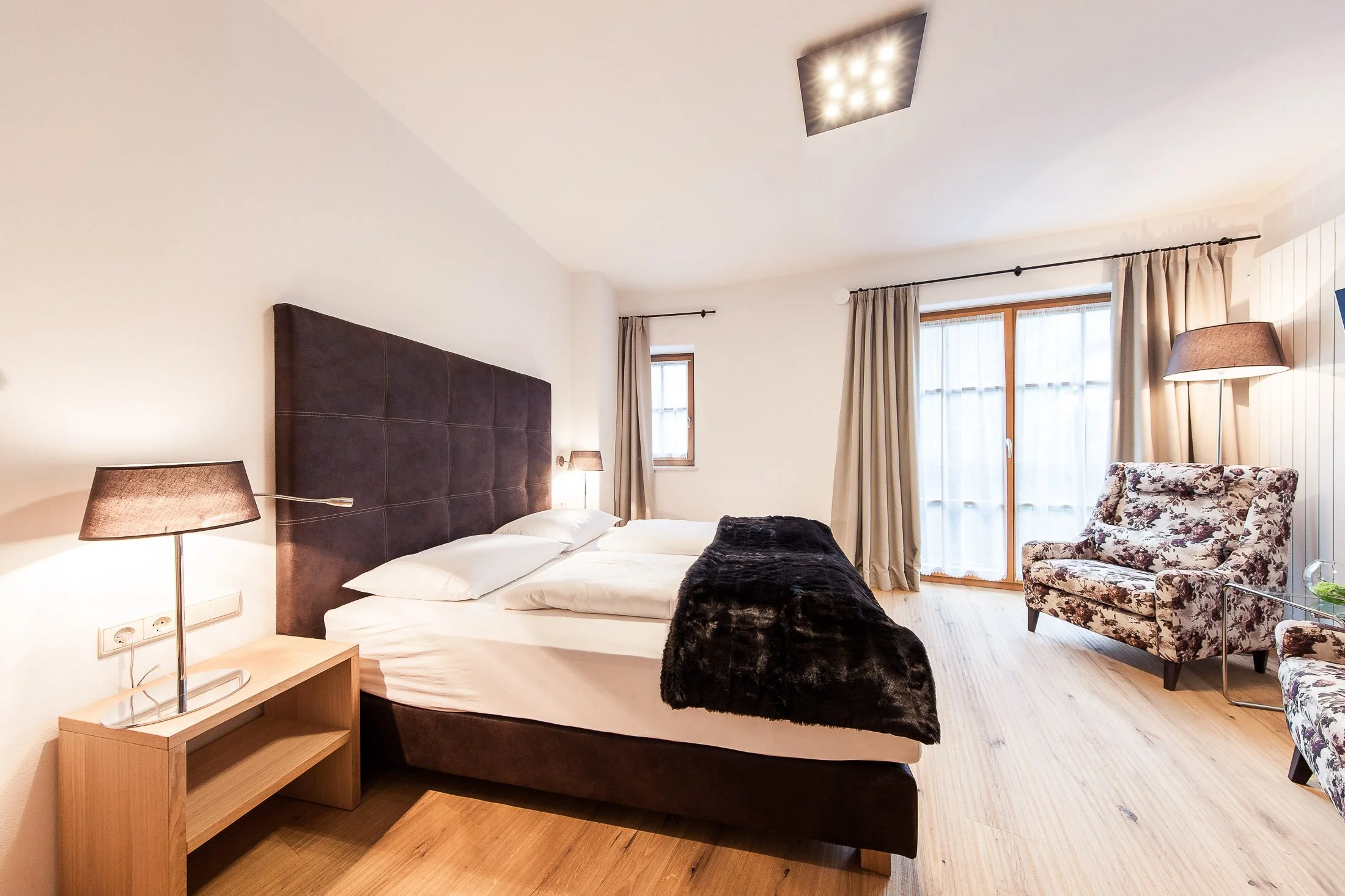 Ein Schlafzimmer mit einem großen Bett, schwarzen Bettdecke, Nachttisch mit Lampe, Fenster mit Vorhängen, Sessel mit Blumenmuster, Stehlampe, Holzboden und moderner Deckenleuchte.
