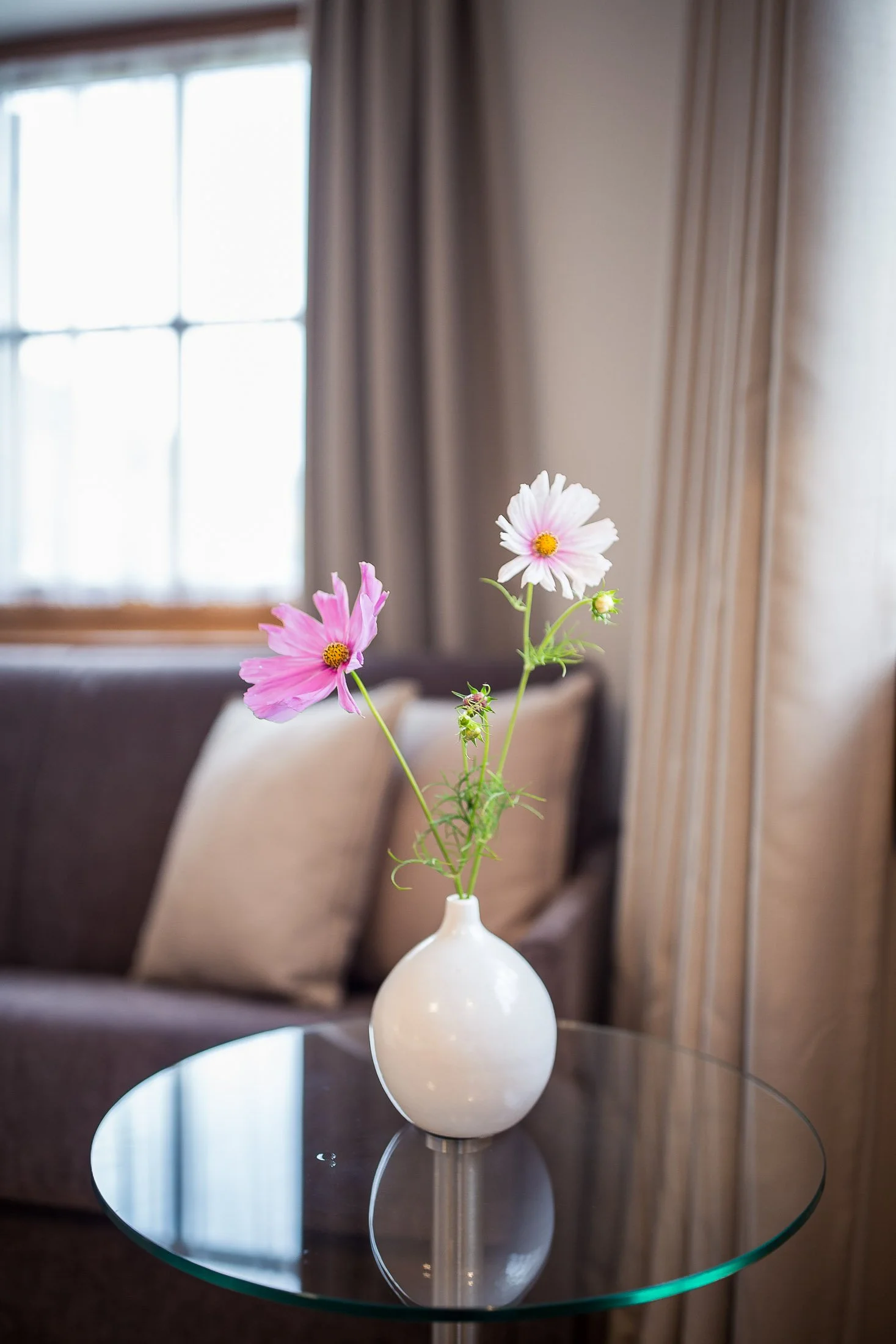 Weiße Vase mit rosa und weißen Blumen auf einem runden Glas-Tisch in einem Wohnzimmer mit großen Fenstern und grauen Vorhängen.