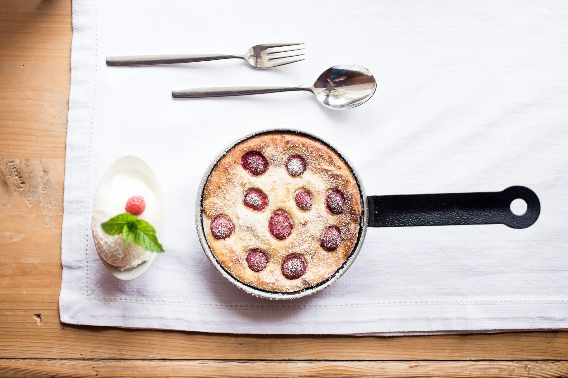 Kleinpfanne mit Kuchen mit roten Beeren, Puderzucker und einer hochwertigen Metallgabel und -löffel, dazu ein kleines Gericht mit Sahne, Minze und Himbeere auf weißem Tuch auf einem Holztisch.