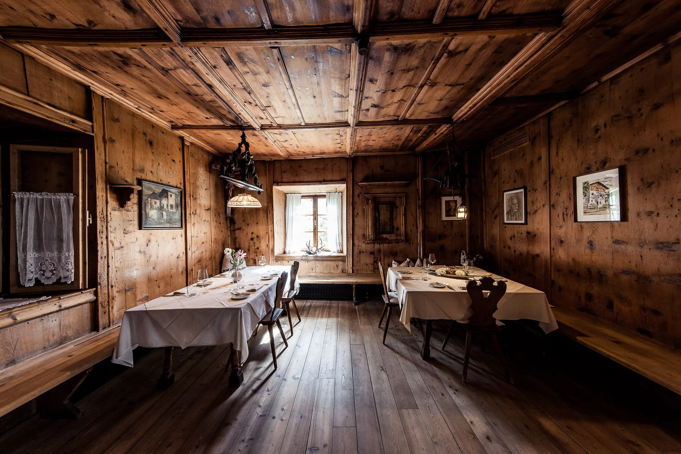 Ein rustikaler Speiseraum mit Holzvertäfelung, bemalten Bildern und gedecktem Tisch mit weißen Tischdecken, Gläsern und Blumen. Ein Fenster lässt natürliches Licht in den Raum.