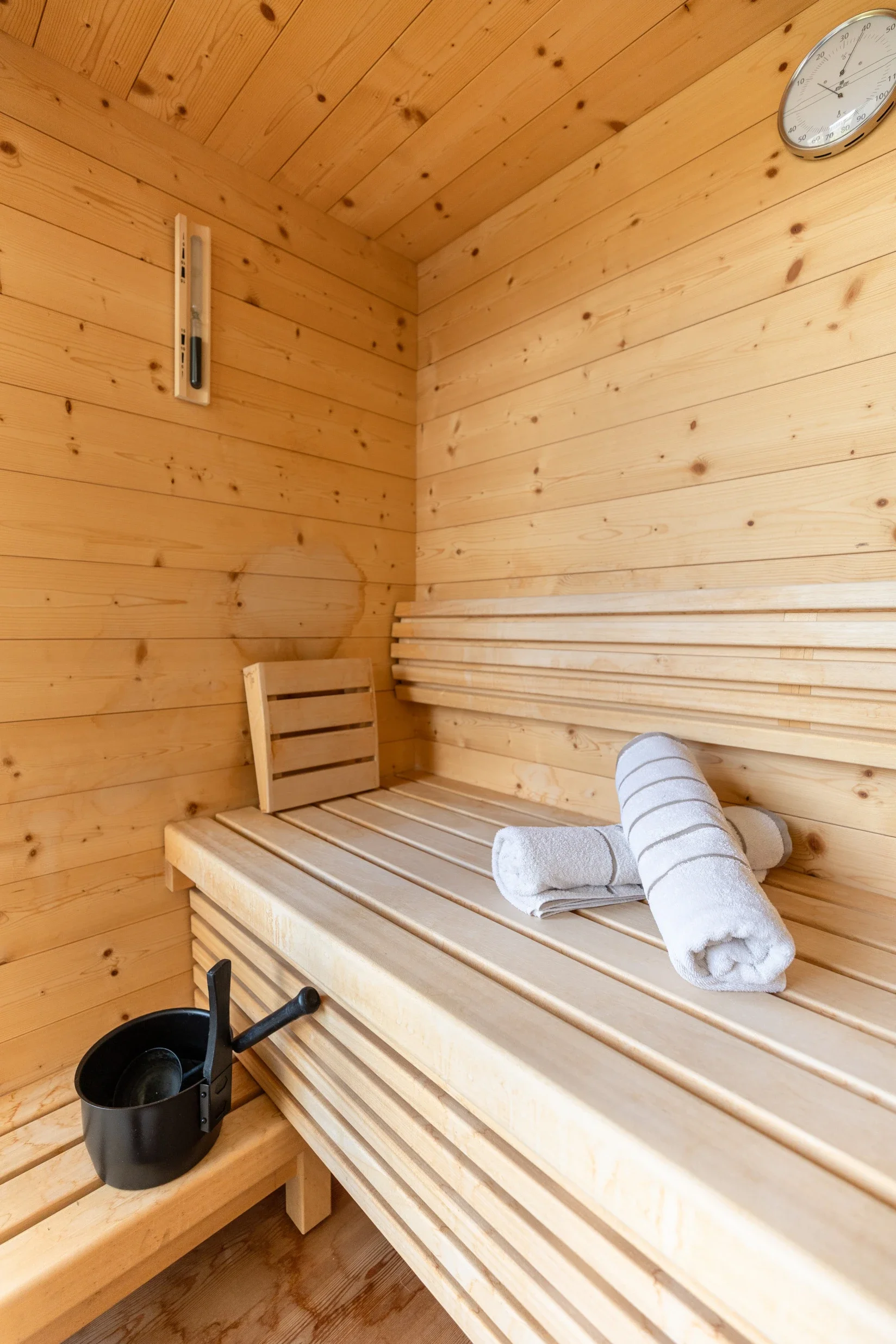 Innenraum einer Holzsauna mit einem Liegestuhl, weißen Handtüchern, einem Saunaofen, einem Thermometer und einer Uhr an der Wand.