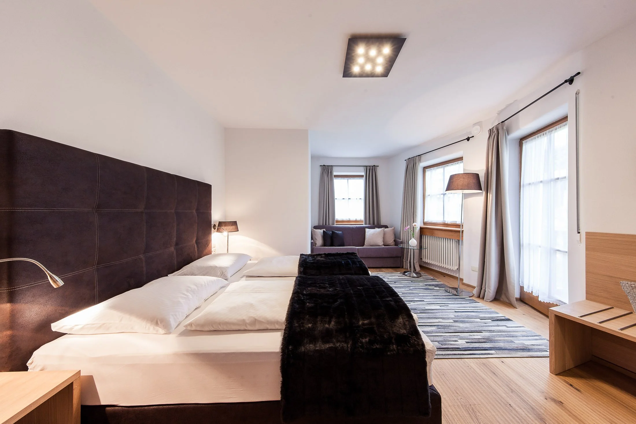 Ein modernes Schlafzimmer mit einem großen Bett, einem hohen braunen Kopfteil, einem Nachttisch mit Leselampe, einem Sofa, großen Fenstern mit Gardinen, einem Stehlampen und einer Teppich im Raum.