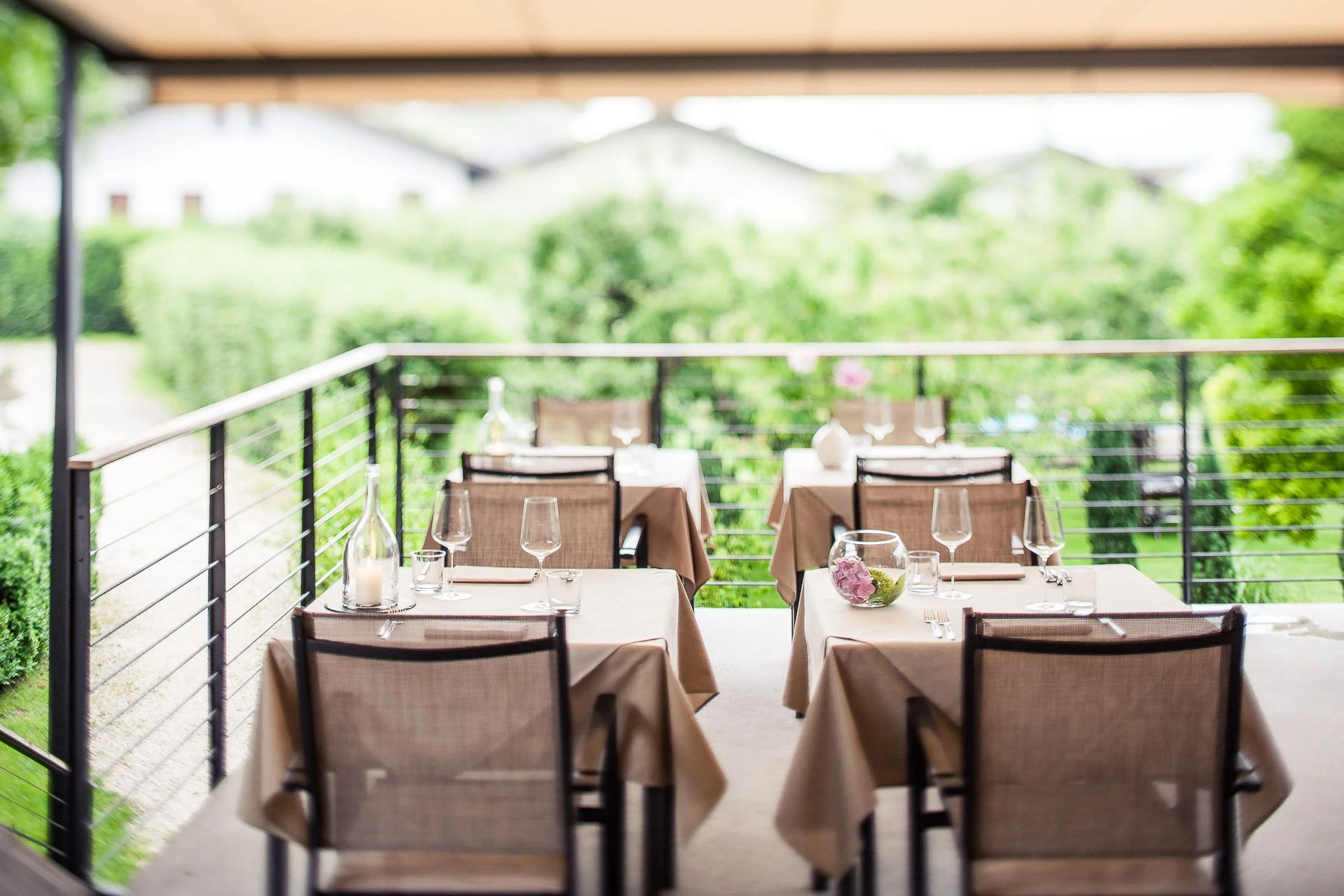 Blick auf einen hübschen, im Freien gelegenen Essbereich auf einer Terrasse mit Tischen, gedecktem Tisch, Gläsern, Kerzen, Blumen und einer grünen, bewaldeten Umgebung.