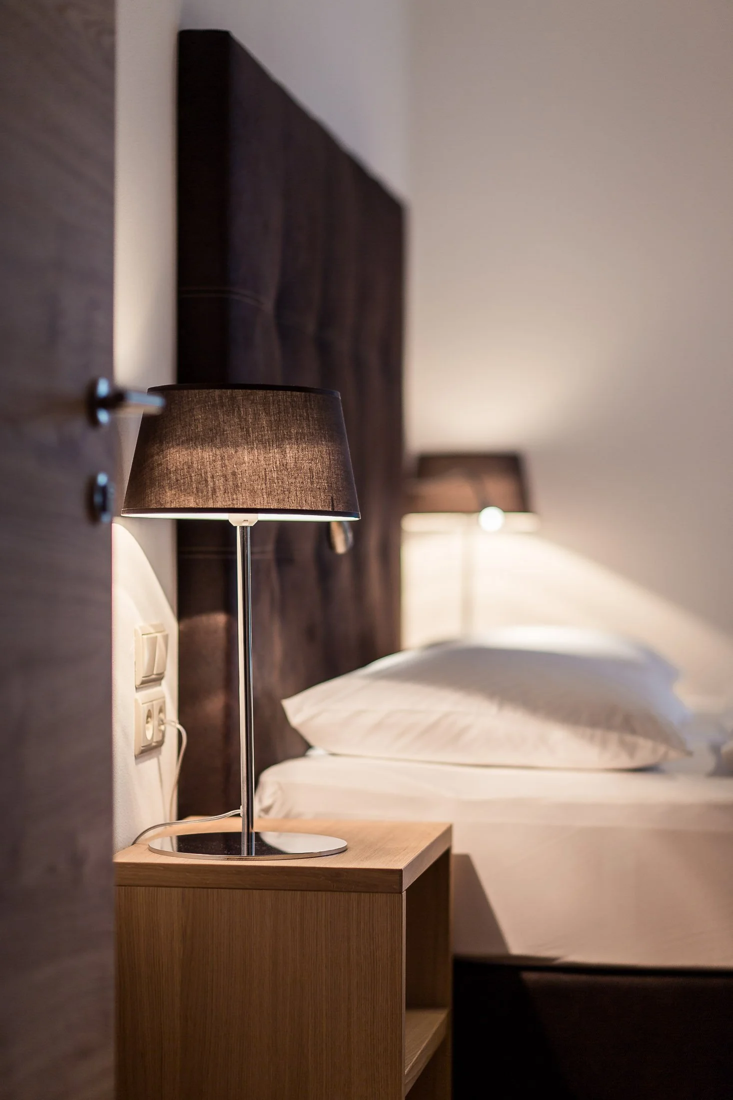 Ein Schlafzimmer mit einem Nachttisch aus Holz, einer Lampe mit grauem Schirm und einem Bett mit weißen Bettlaken und Kissen, dunkelbraunem Kopfende und weißen Wandflächen.