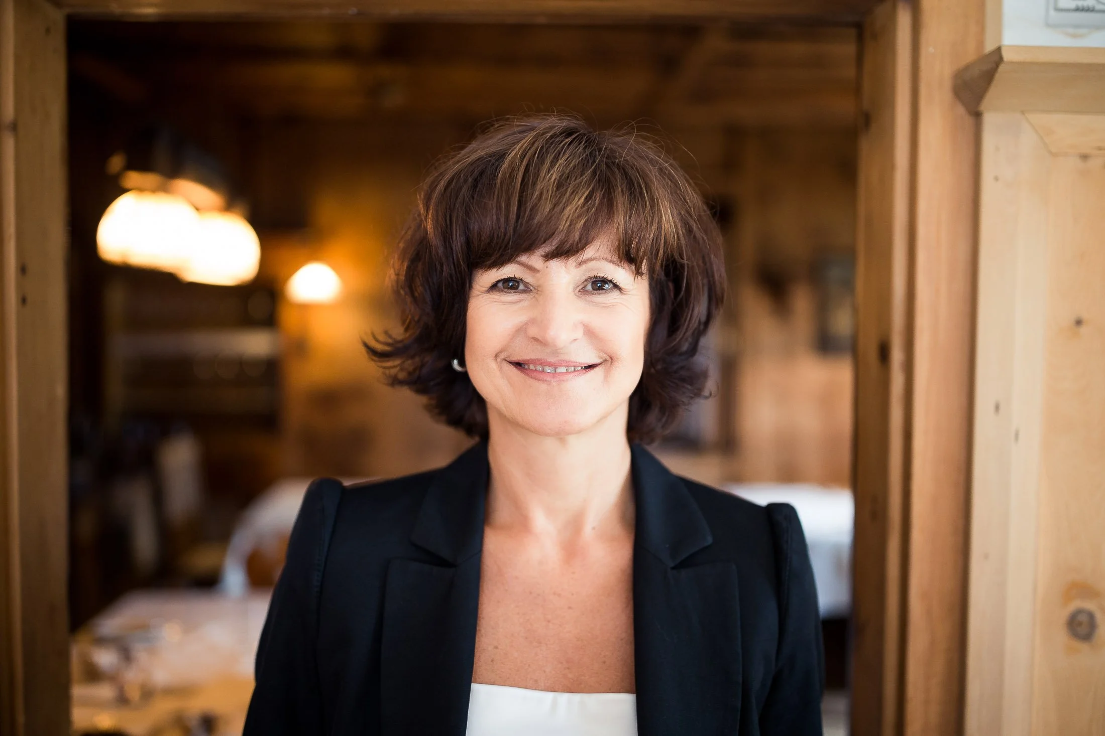 Porträt einer lachenden Frau mit braunen Haaren in einem schwarzen Blazer, die in einem Holzraum steht.