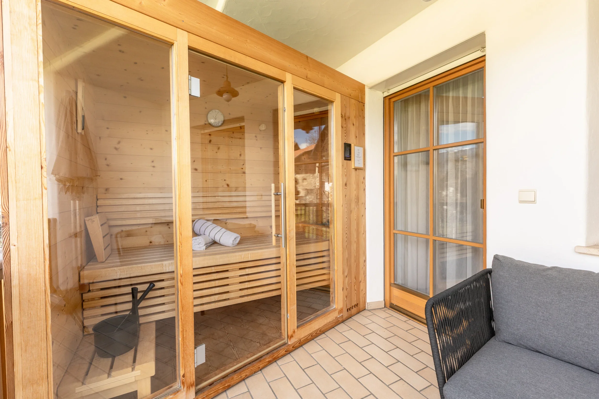 Holzsauna mit Glastür im Außenbereich, neben einer Sitzgelegenheit auf einem Fliesenboden.
