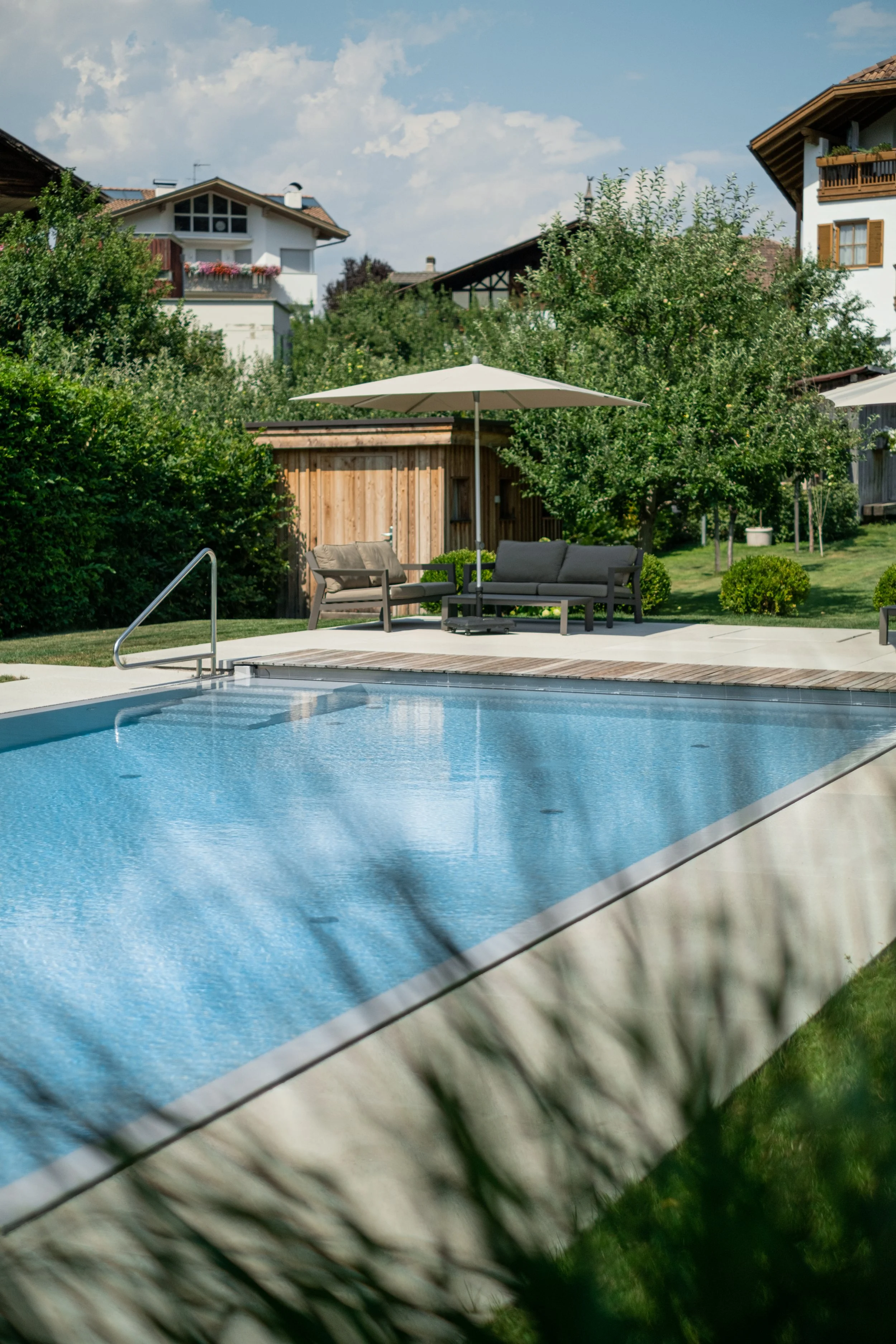 Ein Swimmingpool im Garten mit Teakholzmöbeln, einem Sonnenschirm und umgeben von Bäumen und Häusern bei schönem Wetter.
