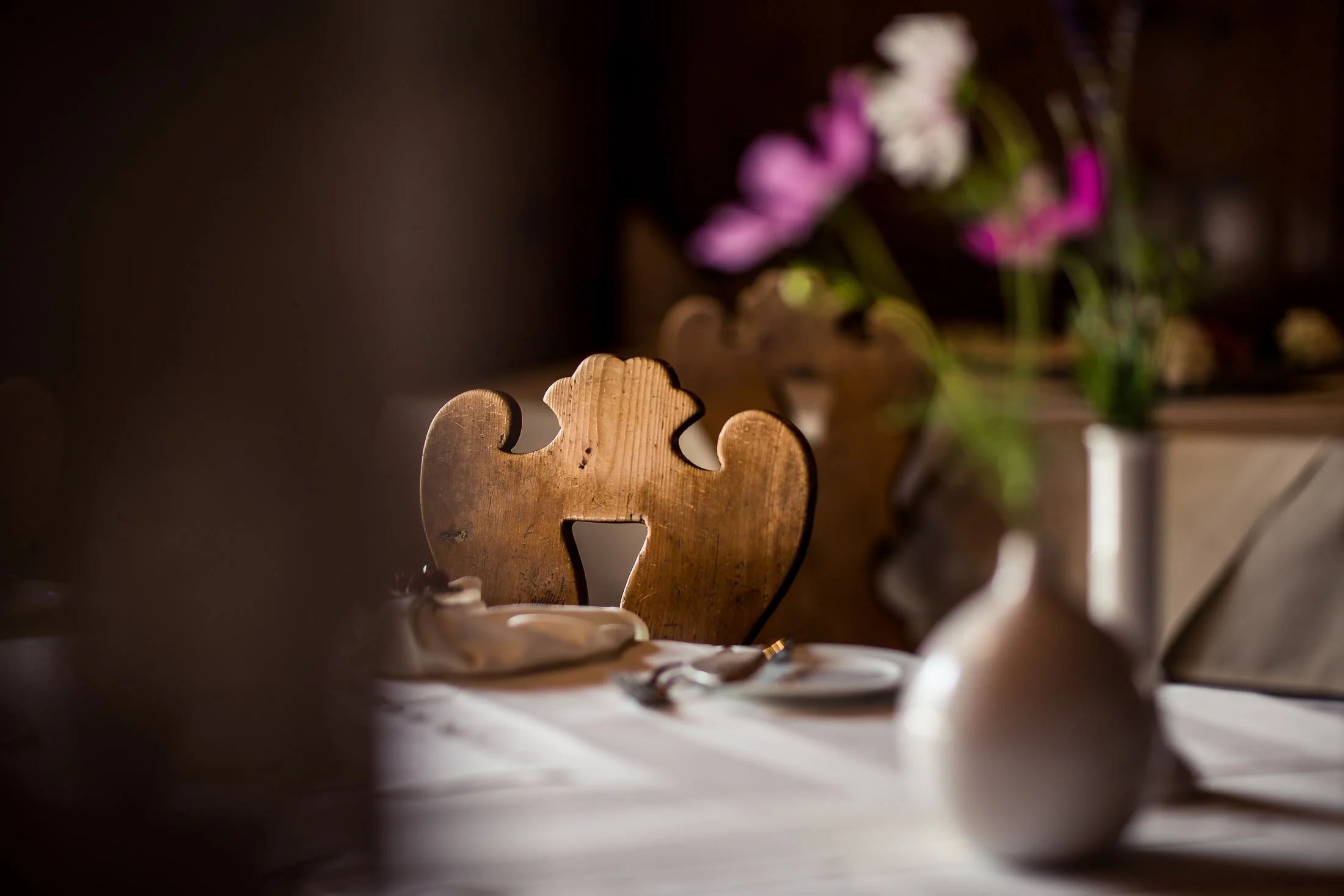 Ein Holzstuhl mit Schnitzereien in einer dunklen Umgebung, im Hintergrund befinden sich bunte Blumen in einer Vase.