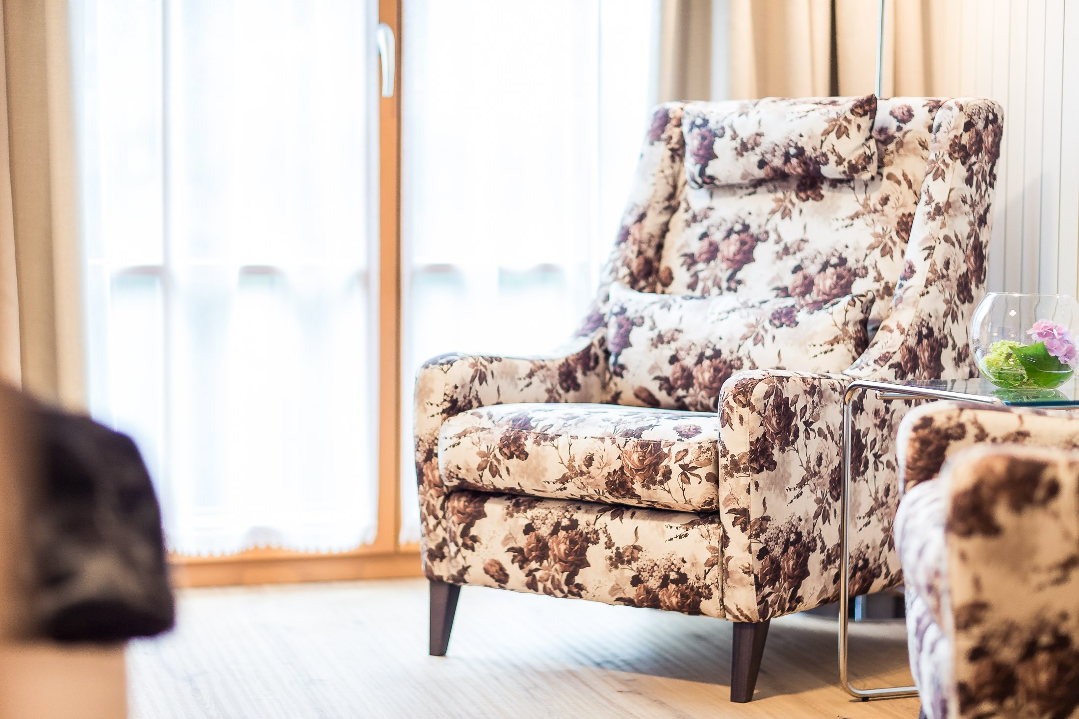 Ein floral gemustertes Sessel in einem Wohnzimmer mit großen Fenstern und Vorhängen.