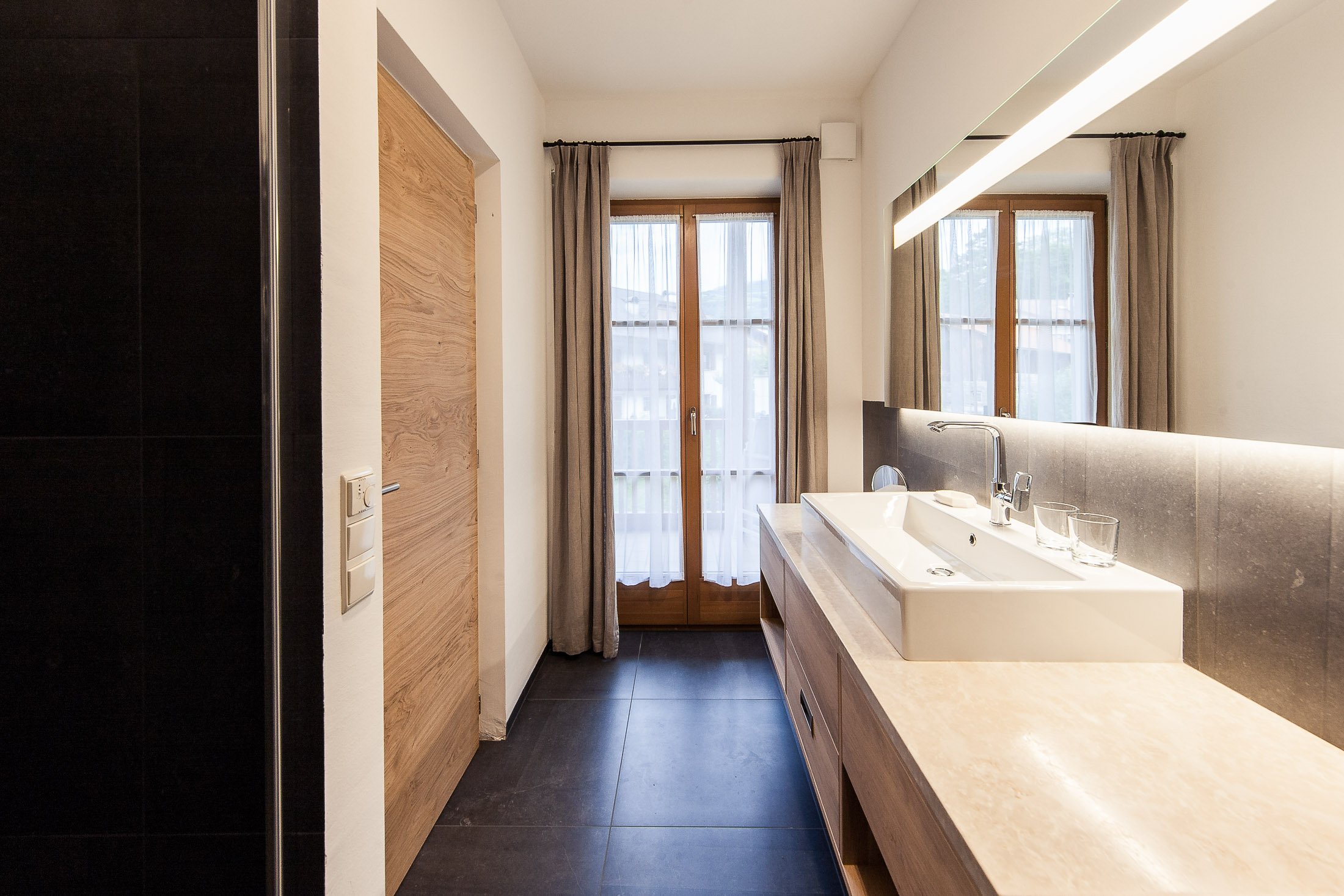 Moderne Küche mit großen Fenstern, weißen Wänden, einer schmalen Theke mit Wasserhahn und zwei Gläsern, beige Vorhängen und dunklen Fliesen.